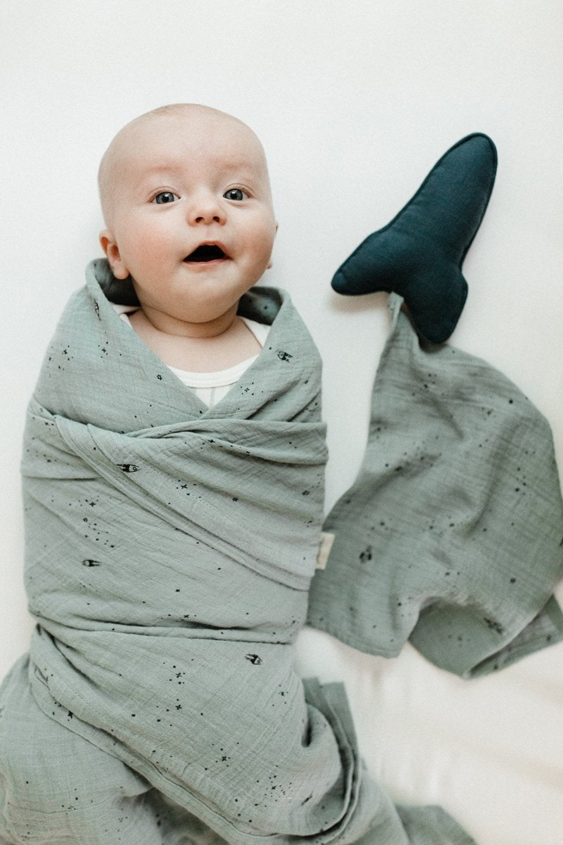 Baby wrapped in Pehr Rocketman Organic Novelty Swaddles beside Pehr Rocketman Lovey. Organic cotton, hand printed. Blue with celestial pattern.