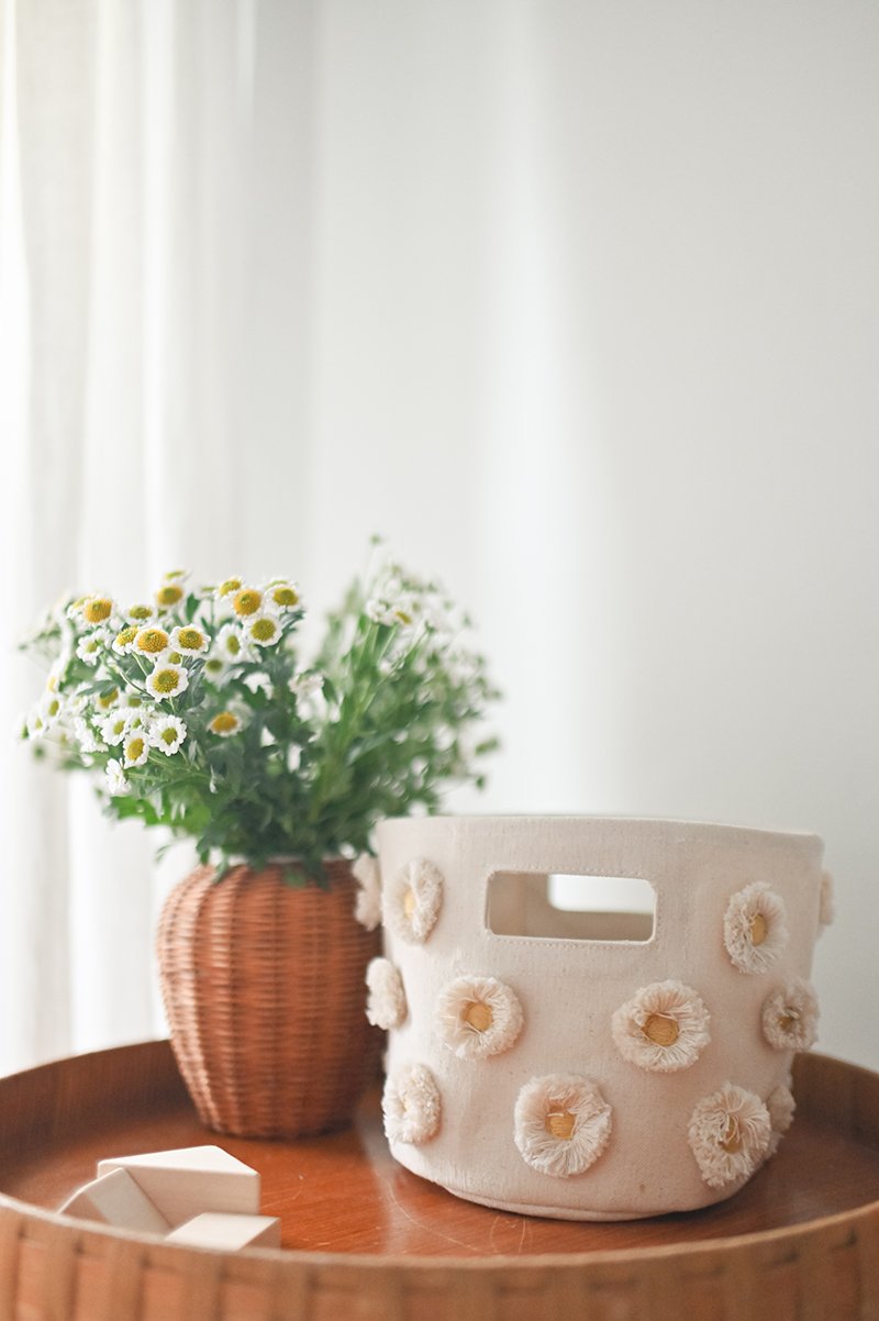 Pehr Daisy Novelty Mini storage bin on table beside potted plant. Screen printed by hand using AZO-Free dyes. Off-white with stitched daisies.