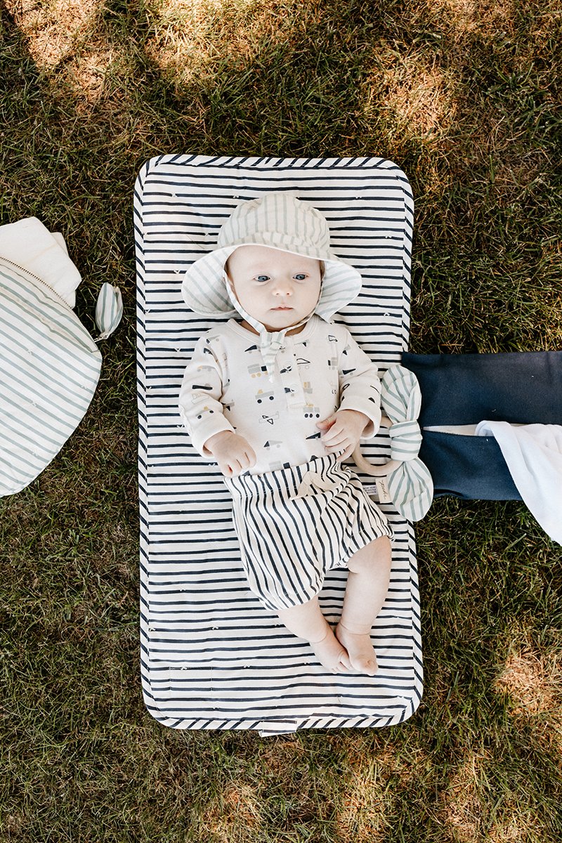 Baby lying Pehr Ink Blue Organic On The Go Travel Change Pad opened up over grass. GOTS Certified Organic Cotton & Dyes. White with dark blue stripes.