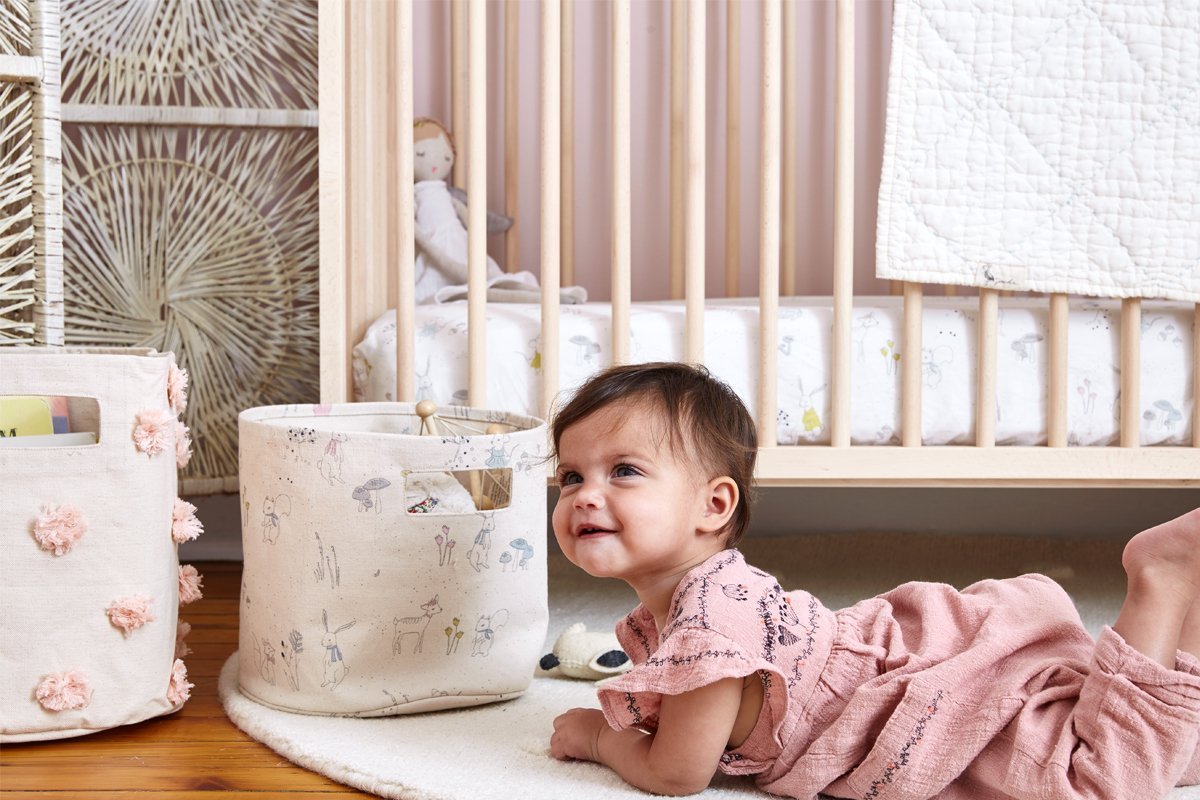 Baby on floor in front of crib with Pehr Magical Forest Organic Crib Sheet. GOTS Certified Organic Cotton. Screen printed by hand using AZO-Free dyes. White with animals.