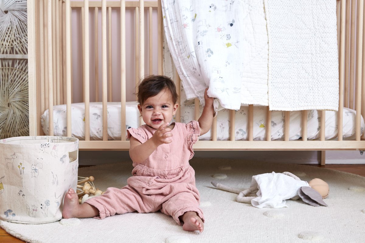 Baby sitting on floor of nursery in front of crib with Pehr Magical Forest Blanket draped over side. Hand printed. White with forest animals and plants on one side, reverse side has coloured specks pattern. Scalloped edging.