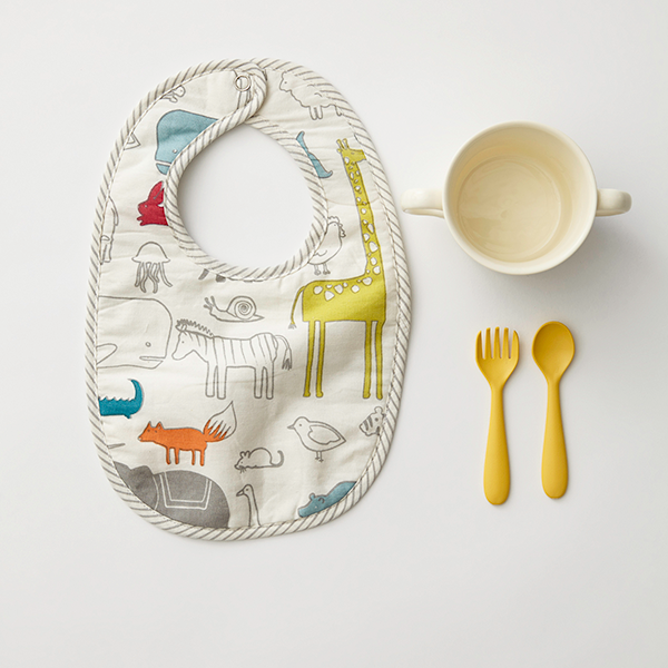 Noah's Ark printed bib from the Pehr Noah's Ark & Painted Dots Bib Set of 2 beside cup, spoon, and fork. Hand printed. White with coloured animals.