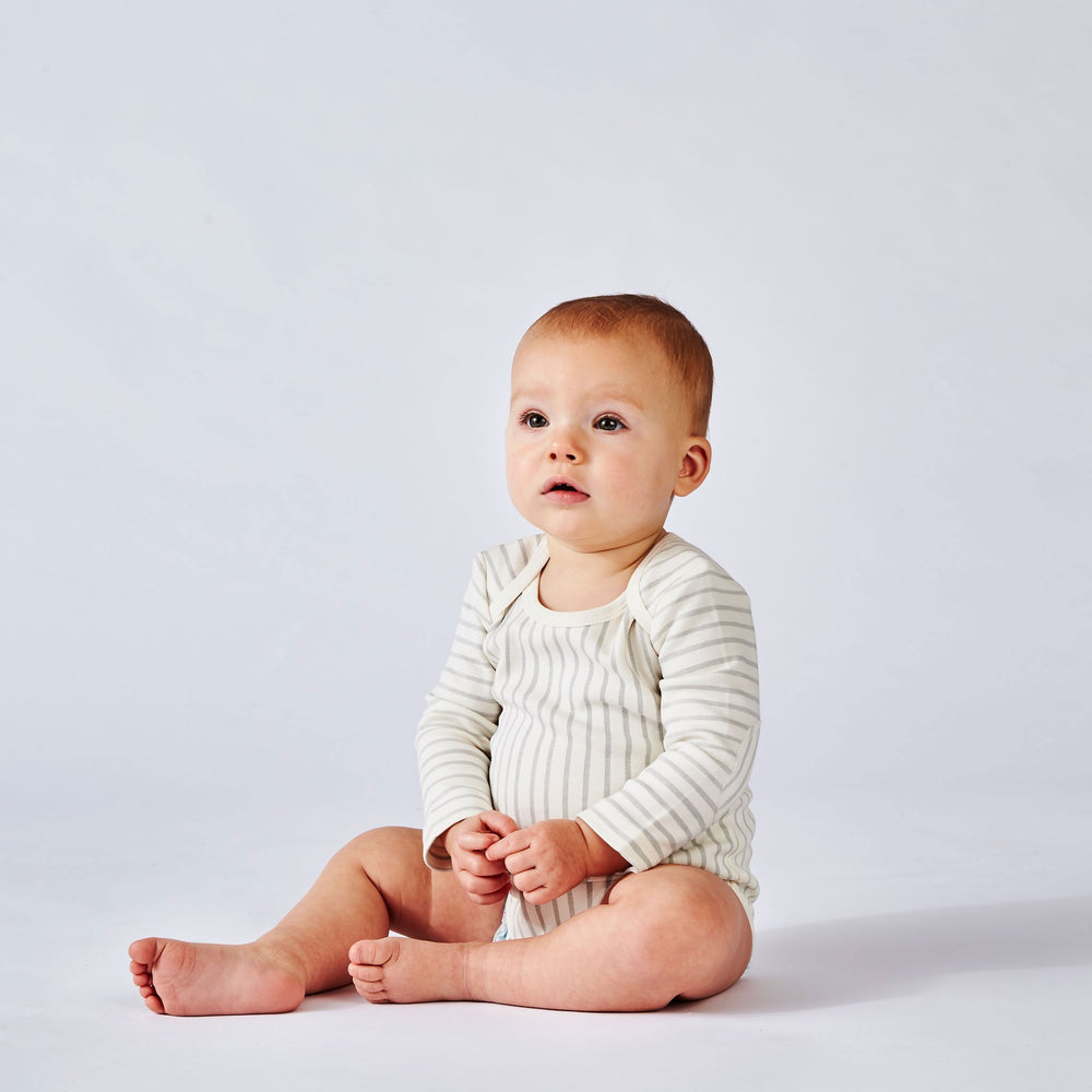 Baby sitting, wearing Pehr Stripes Away Pebble Grey Organic One-Piece, Long Sleeve. GOTS Certified Organic Cotton & Dyes. White with light grey stripes, long sleeve, button closure at bottom.