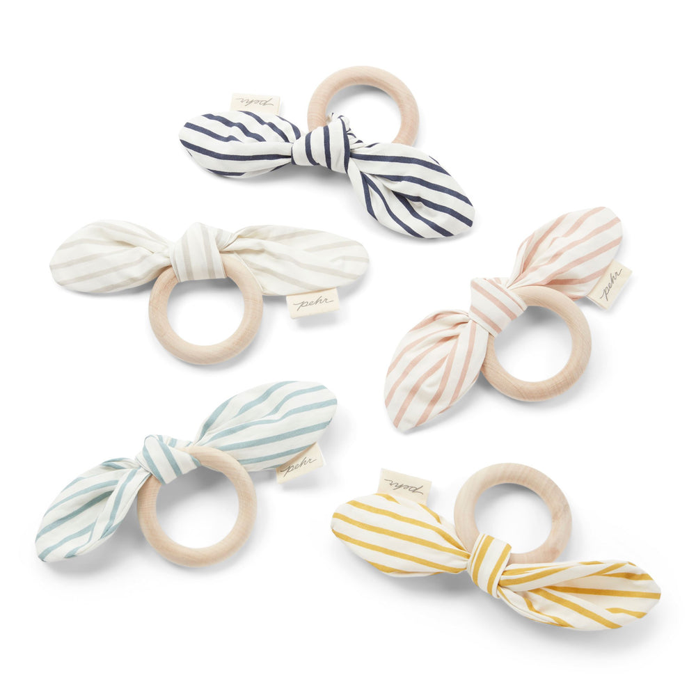 All of the Pehr Organic On the Go Teethers in Rose Pink, Deep Sea, Pebble, Marigold, and Ink Bue. GOTS Certified Organic Cotton & Dyes. White bow with stripes, maple wood ring.