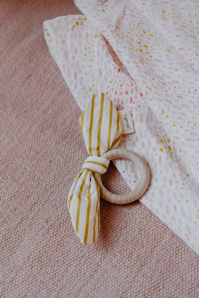 Pehr Marigold Organic On the Go Teether on blanket. GOTS Certified Organic Cotton & Dyes. White bow with gold stripes, maple wood ring.