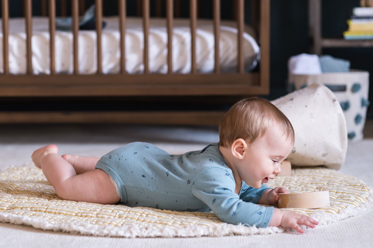 Baby lying on Pehr Celestial Play Mat wearing Pehr Rocketman Organic One-Piece, Long Sleeve. GOTS Certified Organic Cotton & Dyes. Blue with celestial pattern, long sleeve, ruffles on shoulders, button closure at bottom.